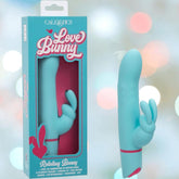 The CalExotics Love Bunny Beaded “G” Bunny, a blue silicone rabbit-shaped vibrator with bunny ear extensions, is shown beside its turquoise packaging with pink "Love Bunny" text. The backdrop features softly blurred pastels.